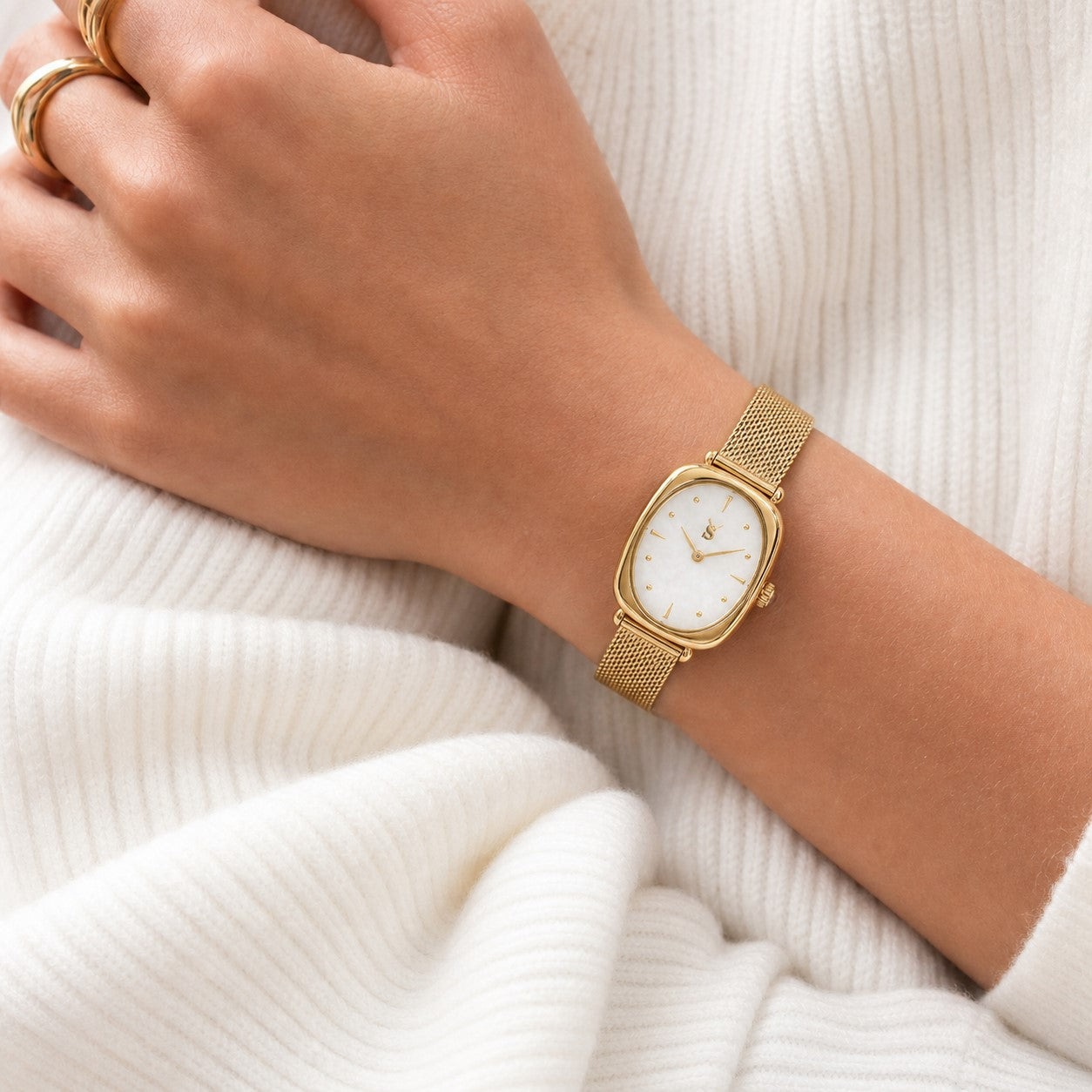 A stylish woman in a cozy white knit sweater wearing the Savwatch vintage-inspired gold mesh bracelet watch and matching gold rings